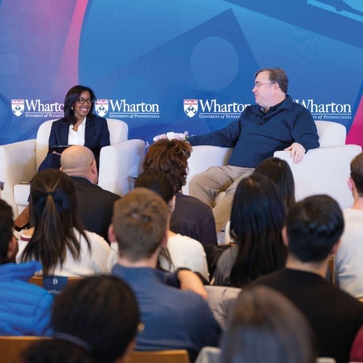 A man and a woman in casual business attire sit on a stage in front of a Wharton-branded backdrop.