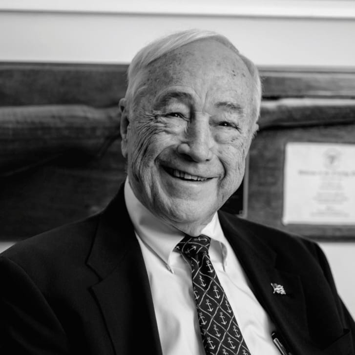 Man in a black suit jacket, white button-down shirt, and a tie with anchors patterned on it.