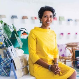 A woman sits in a yellow dress next to a few handbags.