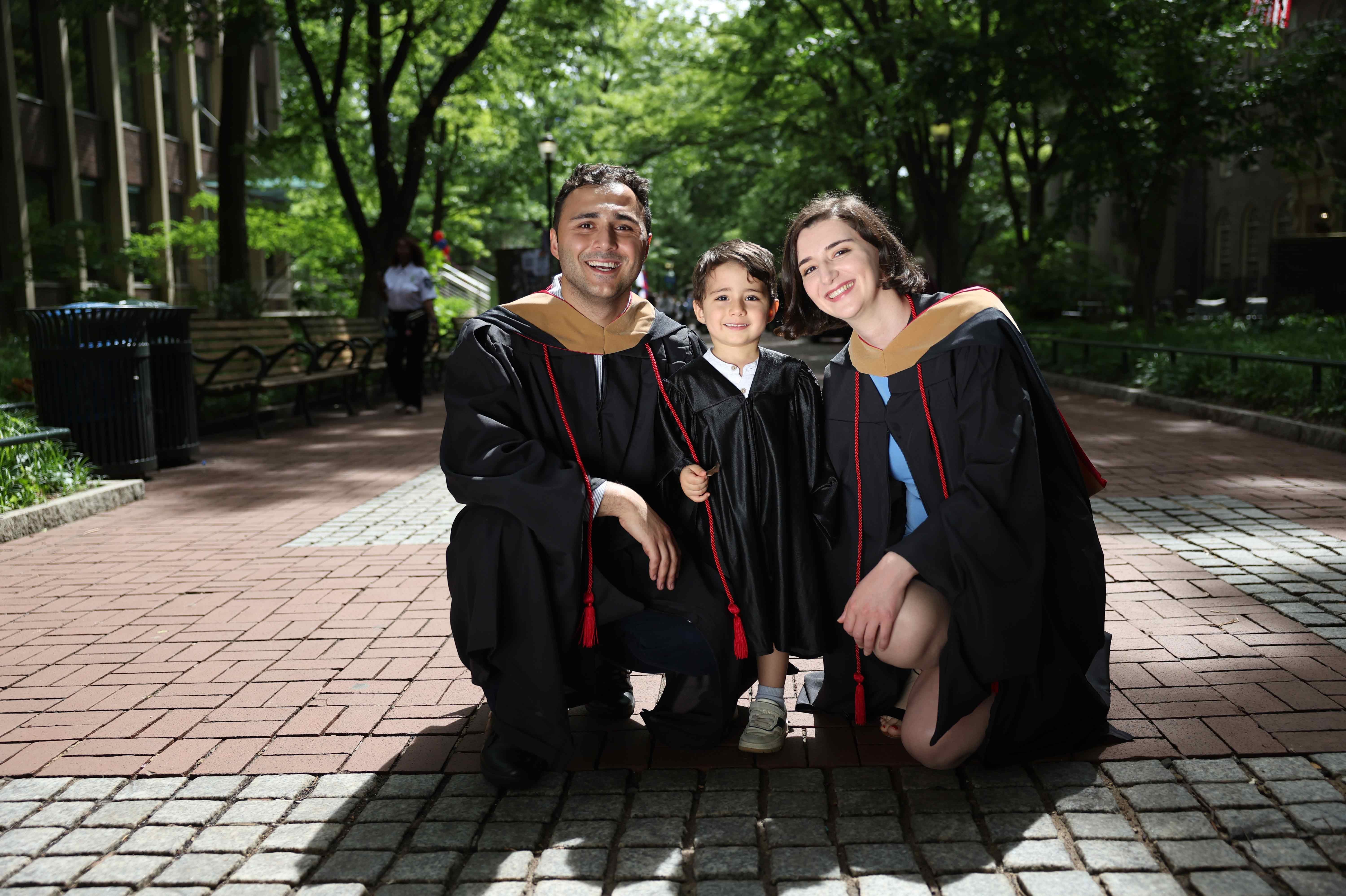 A couple and their son squat down together outdoors on a brick path while wearing graduation gowns.