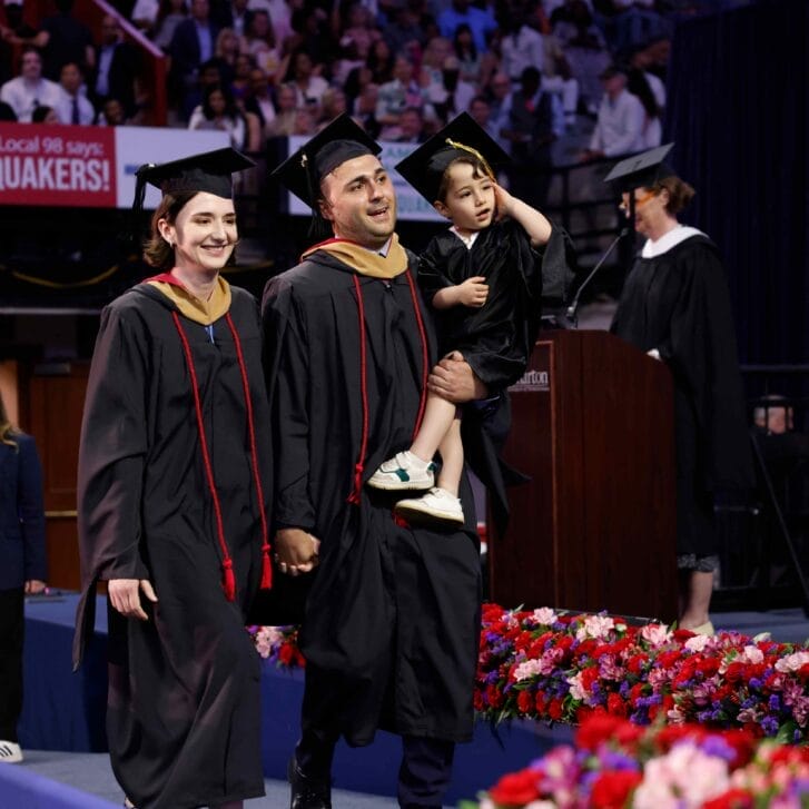 A man carries a boy near his waist while walking next to a woman across a stage during a graduation ceremony.