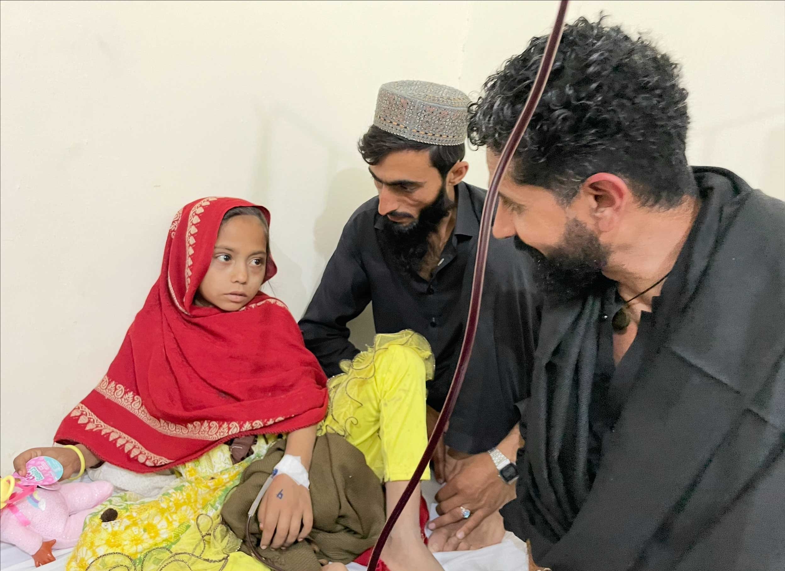 Two men sitting near a young girl with an IV in her hand.