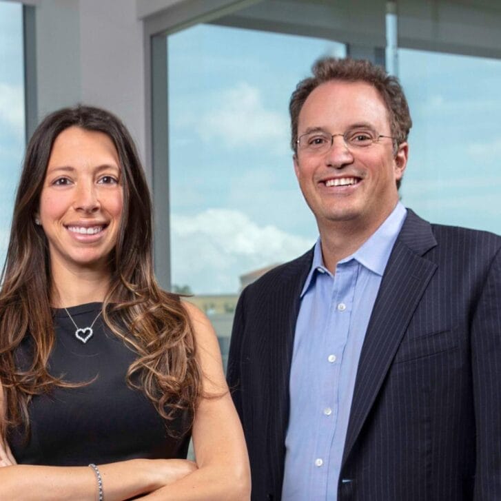 A woman and man in business attire pose for a portrait in front of a set of large windows.