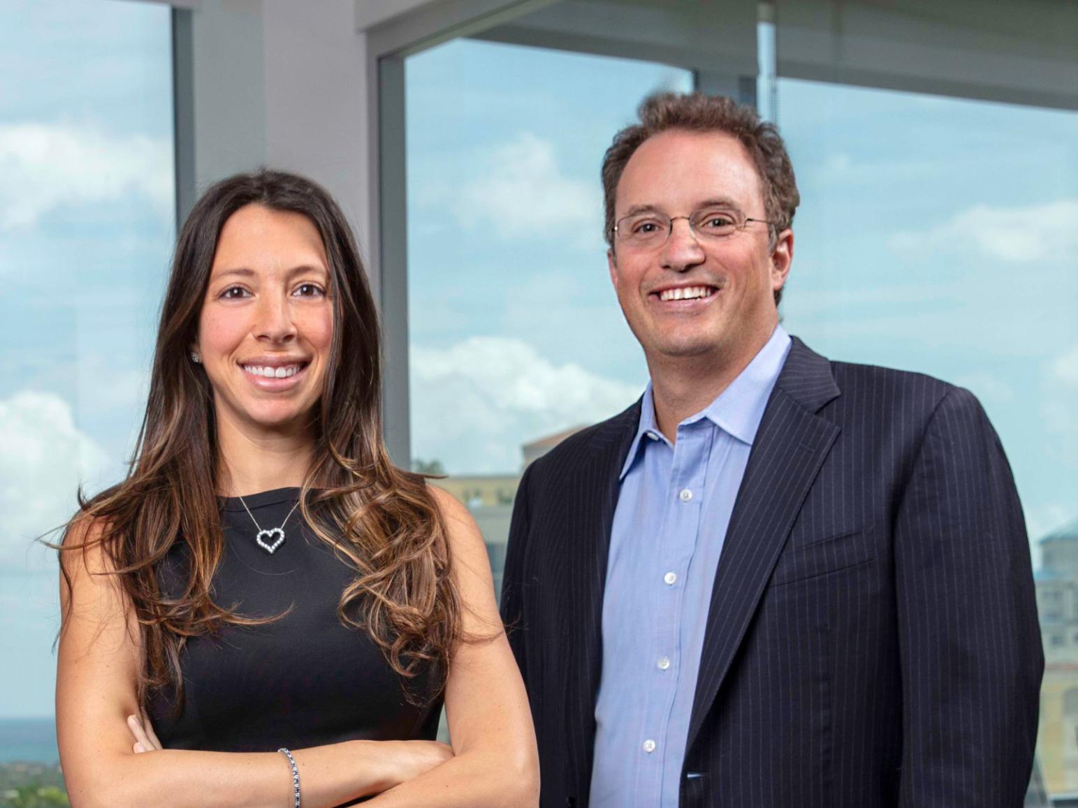 A woman and man in business attire pose for a portrait in front of a set of large windows.