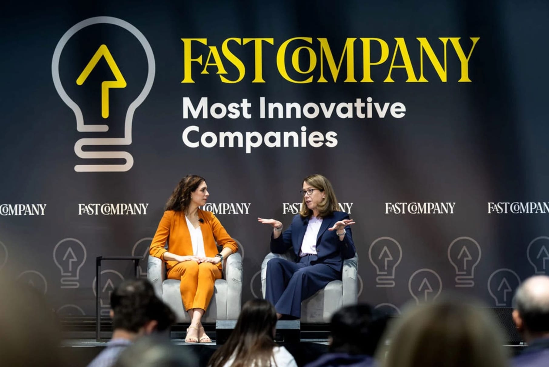 Two women sit on a stage in front of an audience