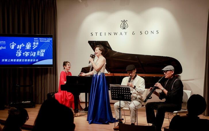 Woman singing into a microphone while standing near a piano and a musical band.