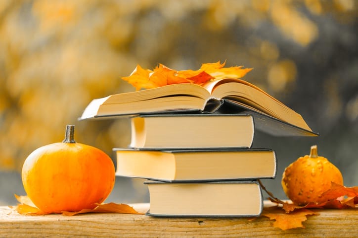 Stack of books surrounded by pumpkins and orange leaves.