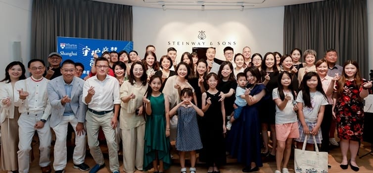 Large group of people standing together in front of a piano and smiling while making hand motions.