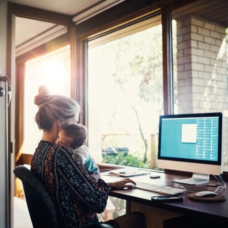 Woman holding a baby while sitting down in front of a desktop computer.