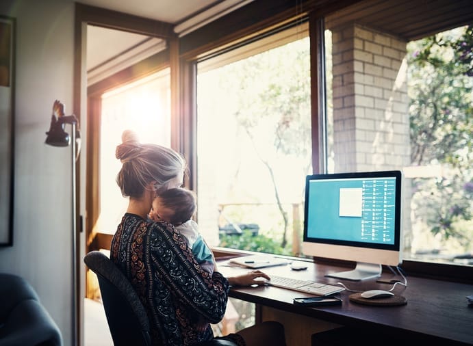 Woman holding a baby while sitting down in front of a desktop computer.
