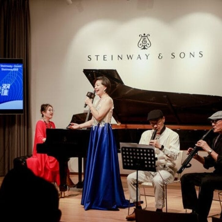 Woman singing into a microphone while standing near a piano and a musical band