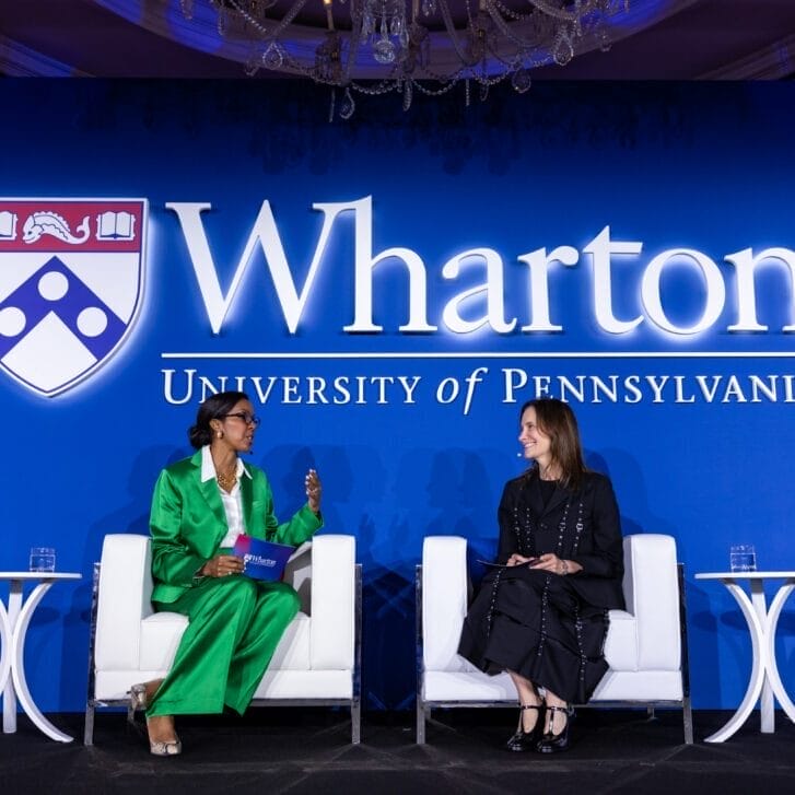 Two women sitting in white chairs on a stage decorated with Wharton - University of Pennsylvania signage