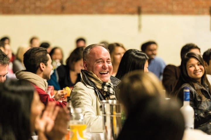 A smiling man seated at a table in a crowded room.