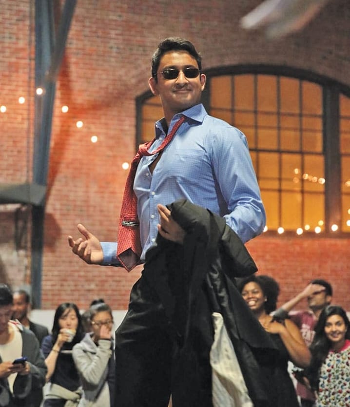 A man in a light blue button-down shirt, sunglasses, and a red tie that is draped loosely around his neck stands on stage holding a suit jacket.