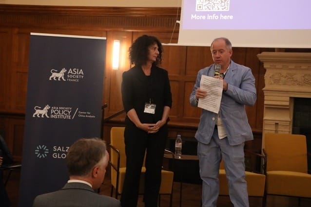 Man speaks into the microphone on a stage while a woman looks on