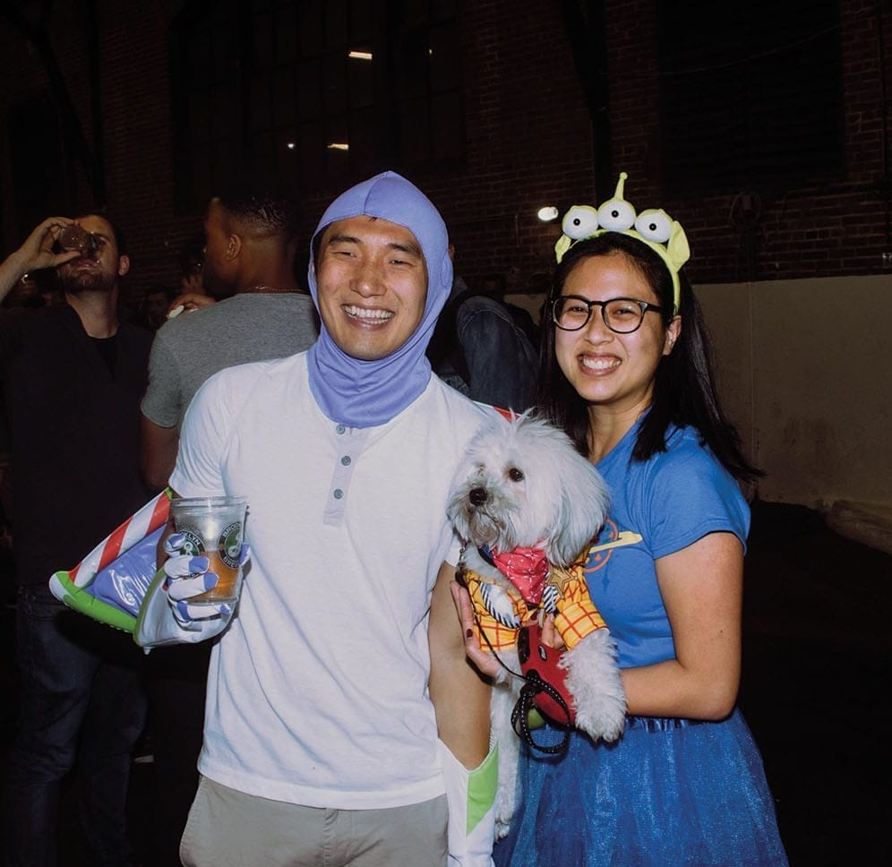 Two people dressed as Toy Story characters pose for a photo with a dog dressed as Woody in one of their hands.
