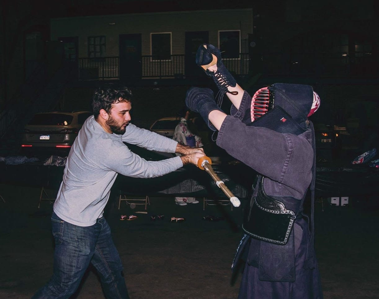 A student wields a shinai in front of a person dressed in a kendo uniform.
