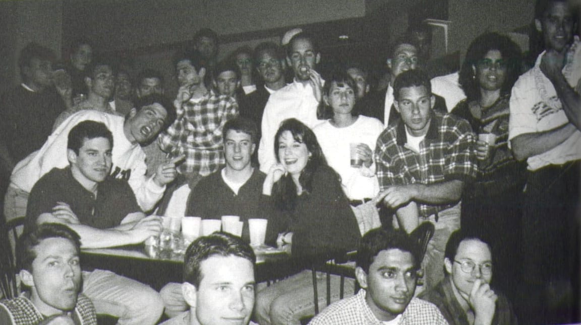 Black-and-white photo of dozens of students crowded around a table, seemingly watching something that is beyond the picture frame.