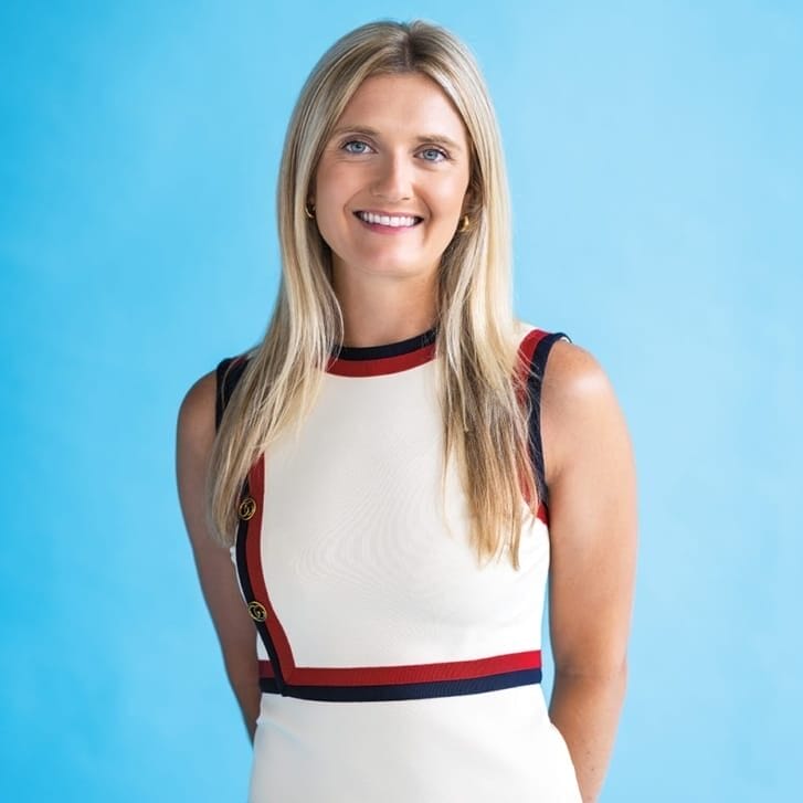 Woman standing with her hands clasped behind her, wearing a white dress with navy and red details.