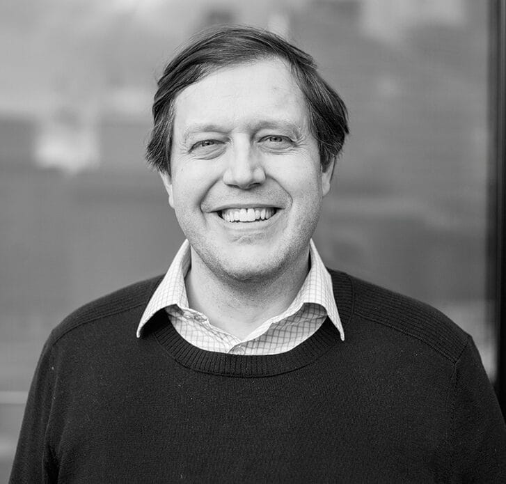 Black-and-white photo of a man smiling and wearing a collared shirt underneath a sweater.