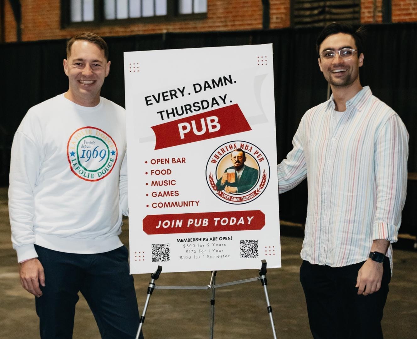 Two men stand on either side of a large sign for Wharton MBA Pub that says "Every Damn Thursday."