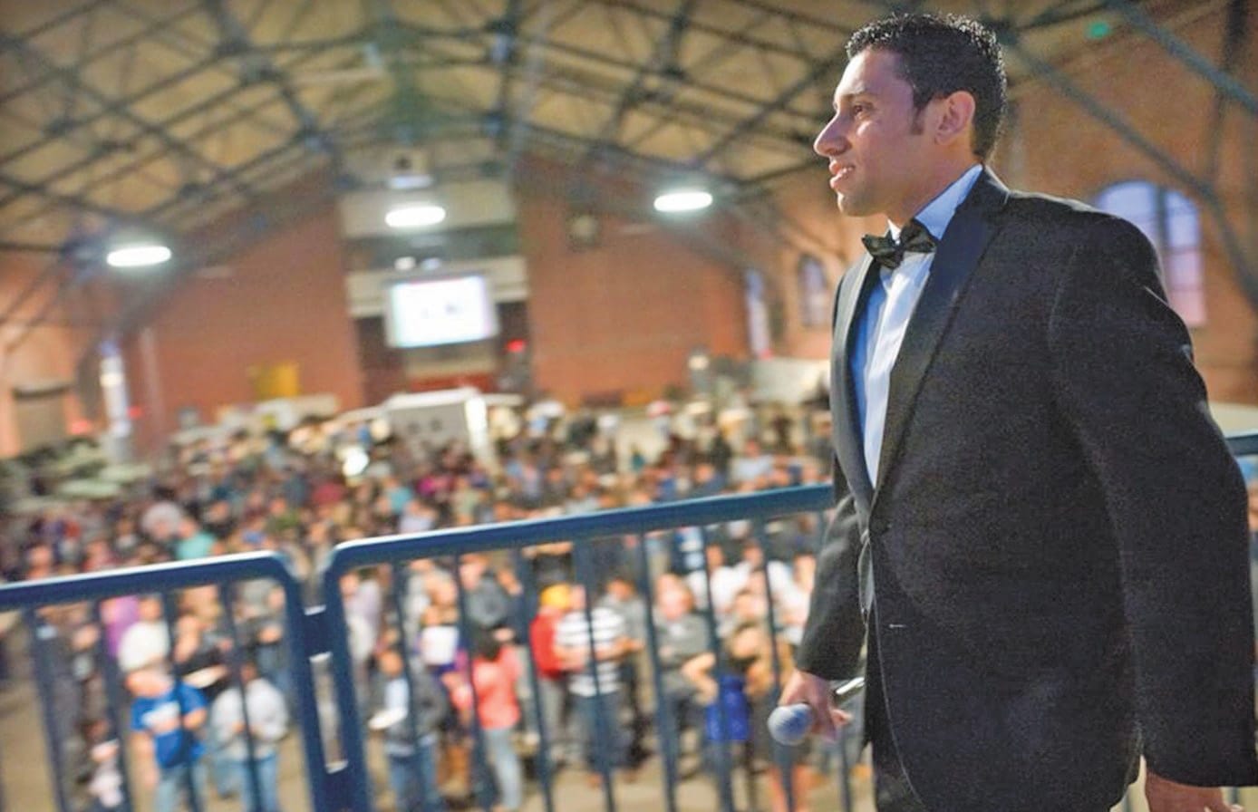 A man in a black suit and bowtie walks across a stage, holding a microphone.