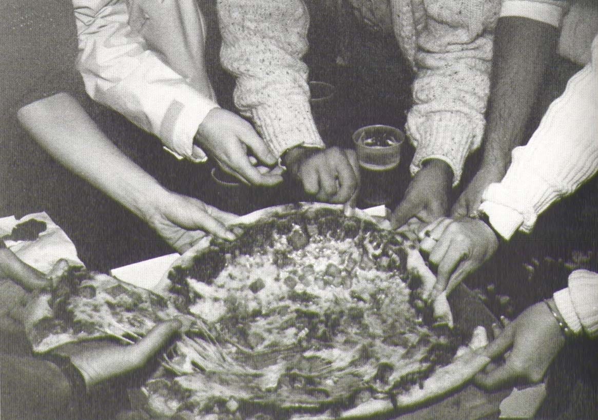 Black-and-white photo of several hands pulling a pizza pie apart.
