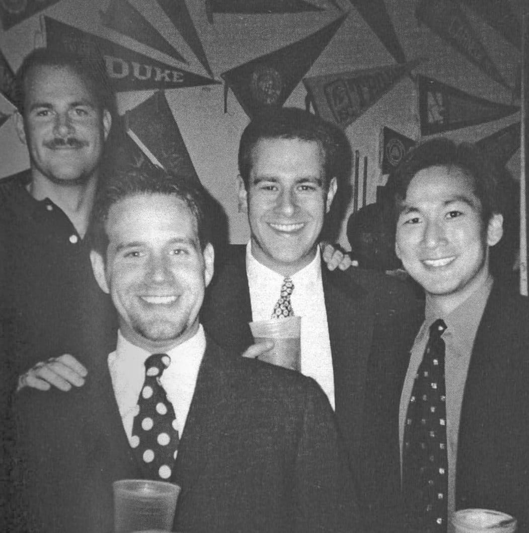 Black-and-white photo of four men in suits and ties smile for a photo.