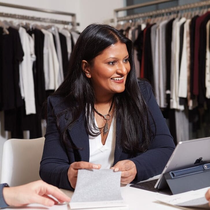 Woman sitting in front of racks of clothing, conversing with others and smiling.