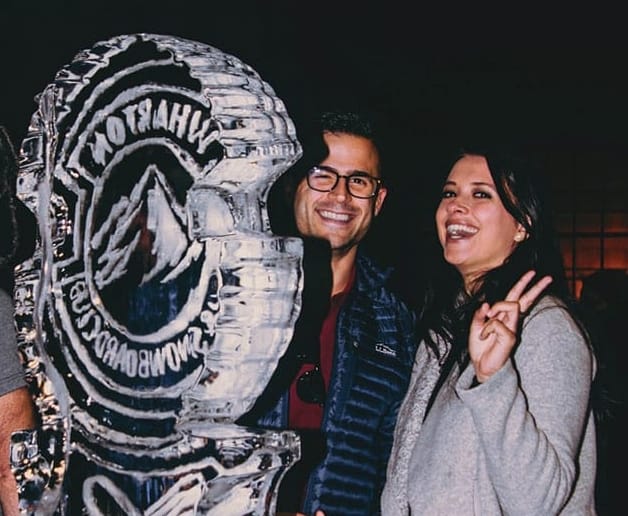 A man and woman flash smiles from their place behind a large ice sculpture.