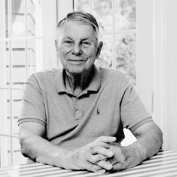 Black-and-white photo of a man sitting in front of a French door with his hands clasped on top of a table.