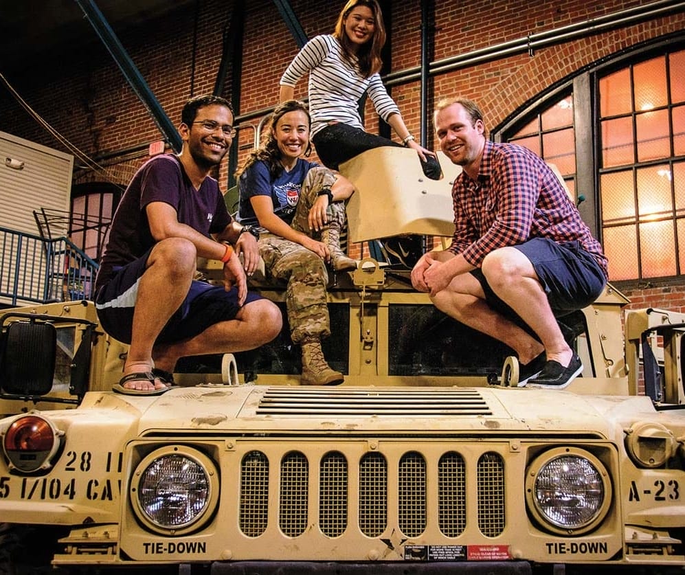 Four people perch atop a military vehicle parked in a drill hall.