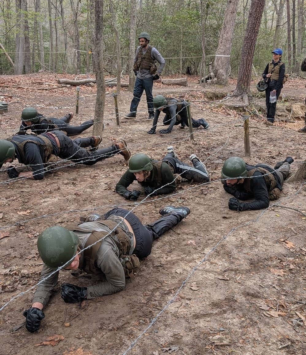 People crawling in a dirt field wearing military garb and hard hats.