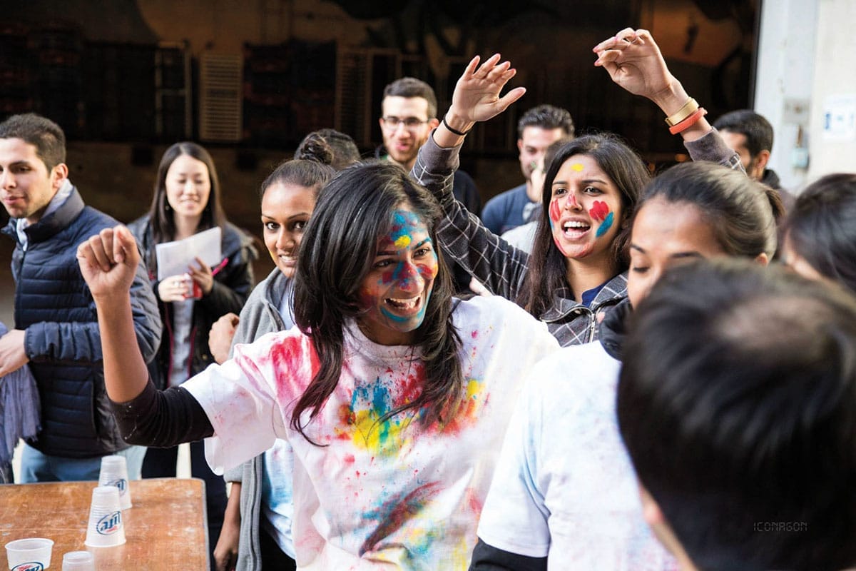 A woman painted in various colors cheers in front of a flip cup table, surrounded by others.