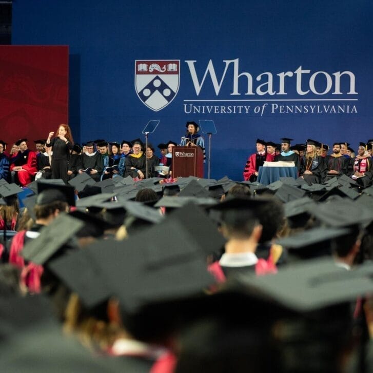 Students wearing black caps and gowns seated in front of a graduation stage.