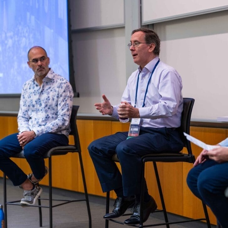 Three men sit on bar stools. The man in the middle gestures while he speaks.