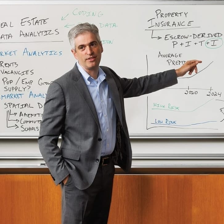 A man in a gray suit and light-blue button-down shirt points to a whiteboard with notes all over it.