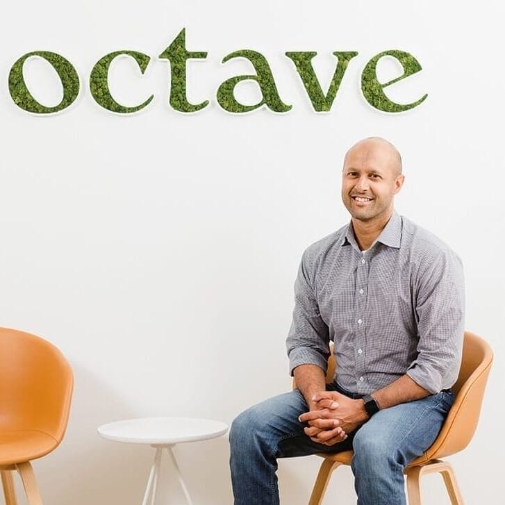 Sandeep Acharya in jeans and a button-down shirt seated in front of a wall with an Octave logo on it.