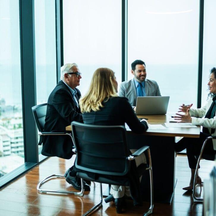 Executives smile at each other and make positive gestures while sitting around a board room table in a glass-walled skyscraper