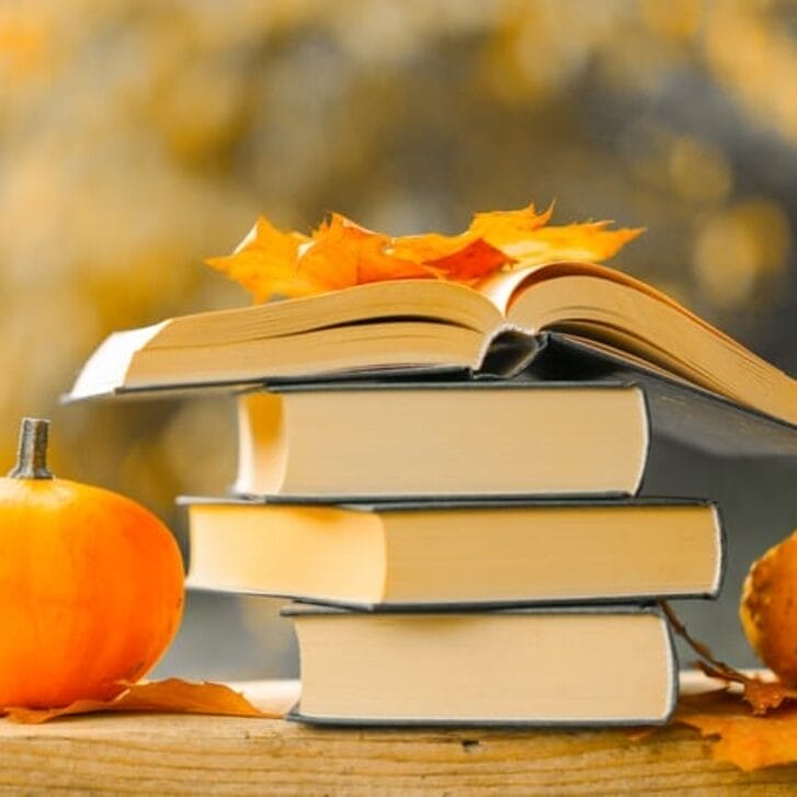 Stack of books surrounded by pumpkins and orange leaves.