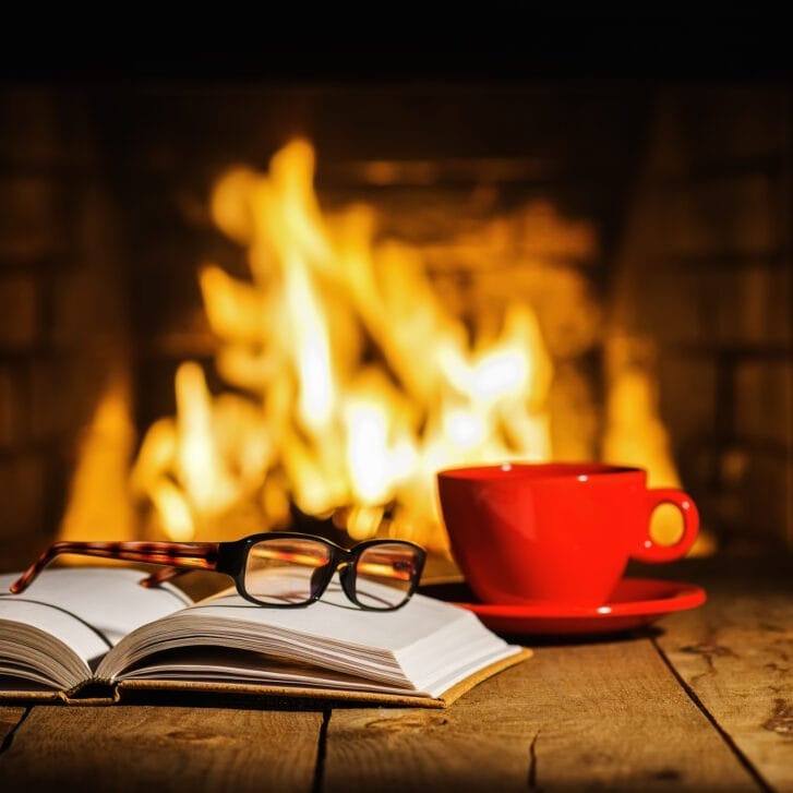 Red cup of coffee or tea, glasses, and old book on wooden table near fireplace.