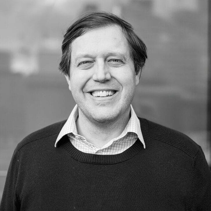 Black-and-white photo of a man smiling and wearing a collared shirt underneath a sweater.