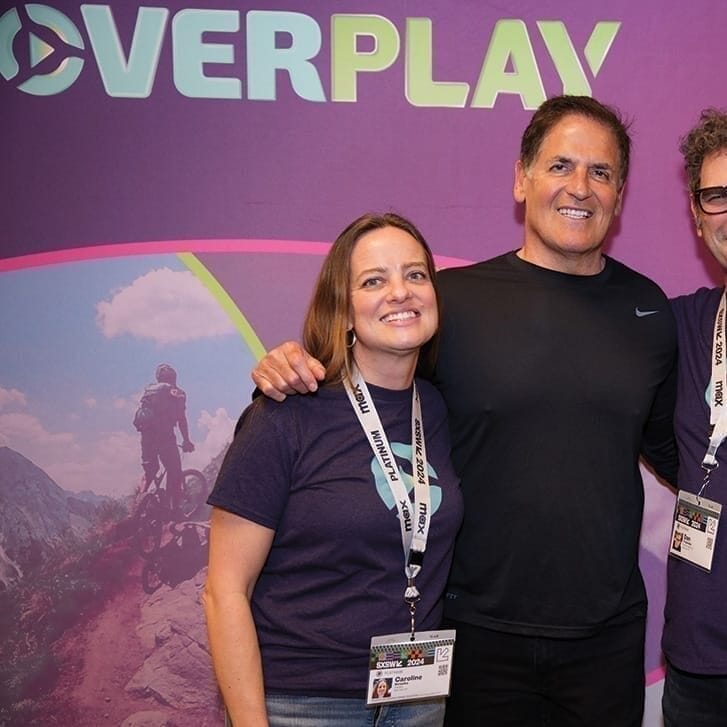Three people pose for a photo together against a backdrop with imagery for the startup Overplay on it.