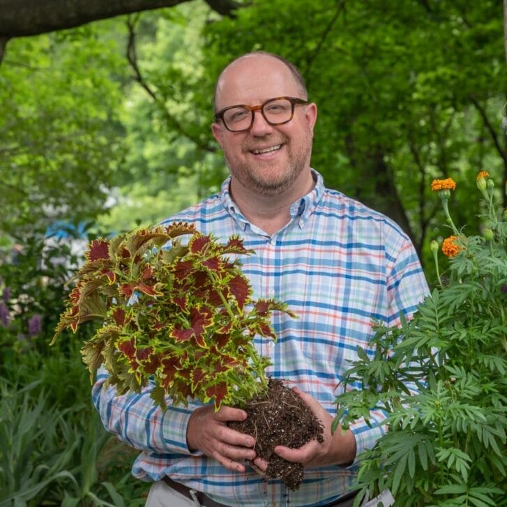 Man in a patterned button-down shirt and glasses smiles and hold an unpotted plant.