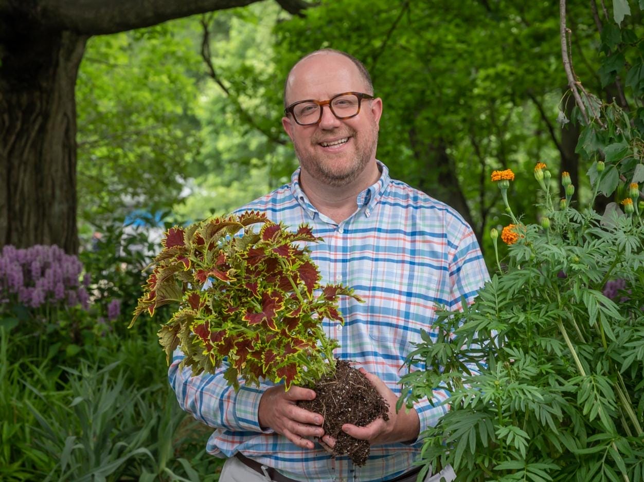 Man in a patterned button-down shirt and glasses smiles and hold an unpotted plant.