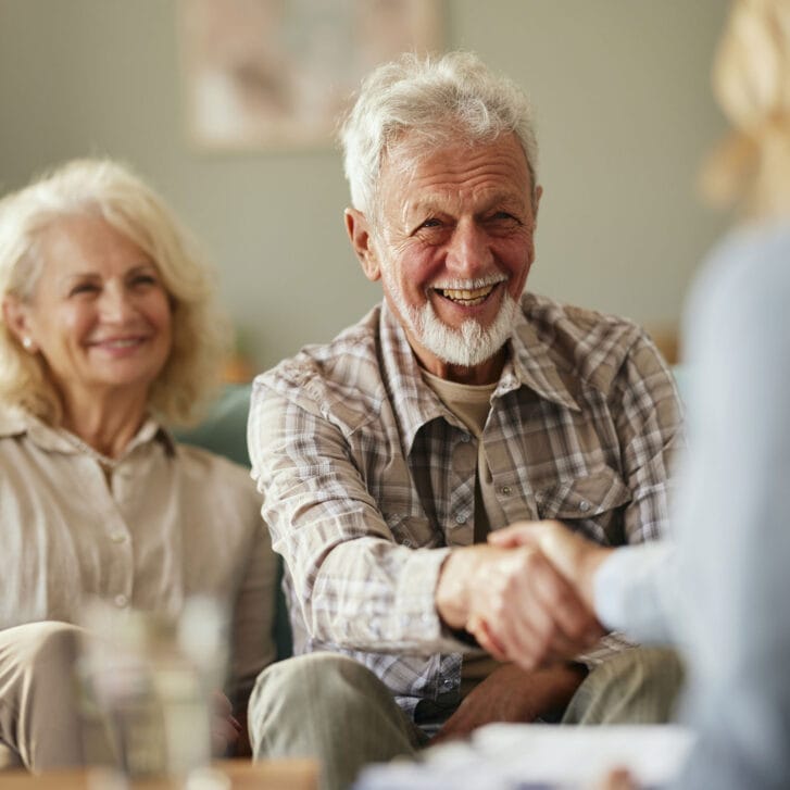 Gray-haired man shaking hands with a woman as his wife sits nearby