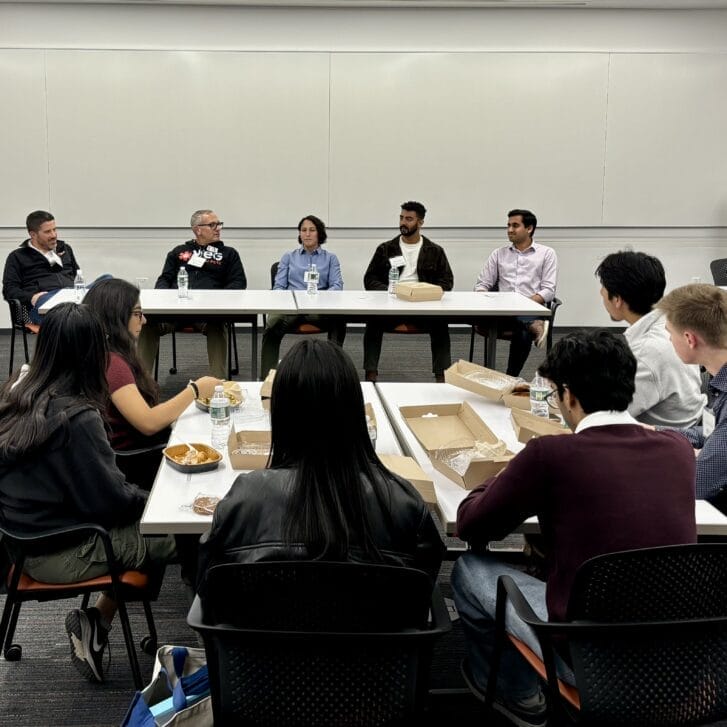 People engaged in a discussion at a long table, as other people eating their lunches look on from another table.