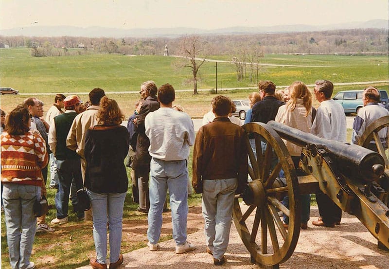 Dozens of people stand outside in front of a cannon looking out over a grassy field.