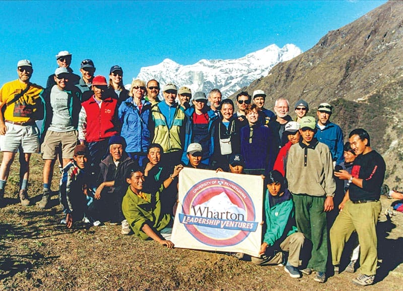 More than two dozen people stand together, with a few people in front holding a sign that says Wharton Leadership Ventures. A snow-capped mountain peak looms in the background.