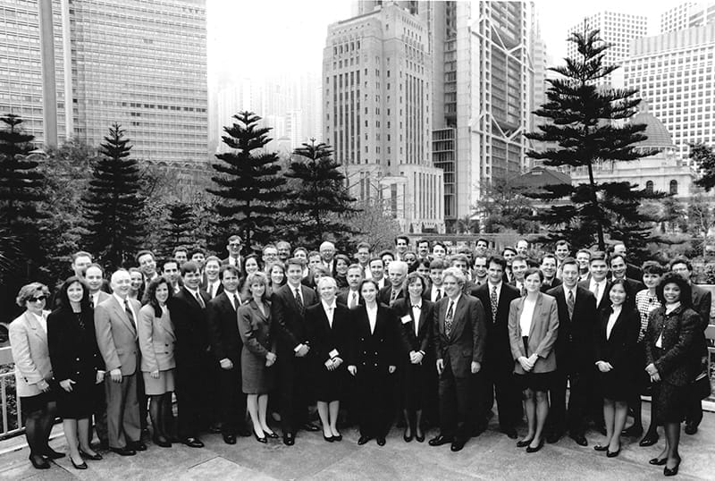Dozens of people in formal business attire pose for a photo in front of tall buildings.